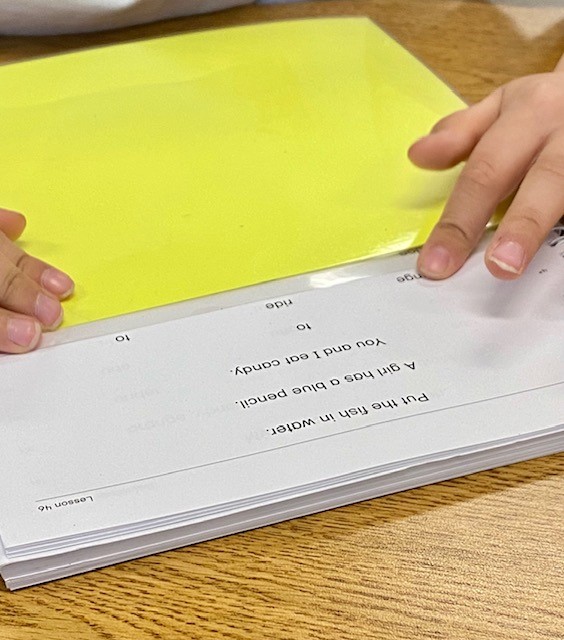 A white spiral-bound book is shown on a table, with a child's hands holding a piece of yellow cardstock to cover some of the lines in the book. The child is pointing to a word on the page.