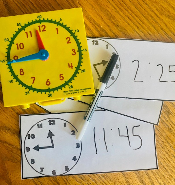 Task cards with analog clocks are shown. the times are written 11:45 and 2:25 are written with a dry erase marker. A small, yellow plastic clock shows the time 11:45.