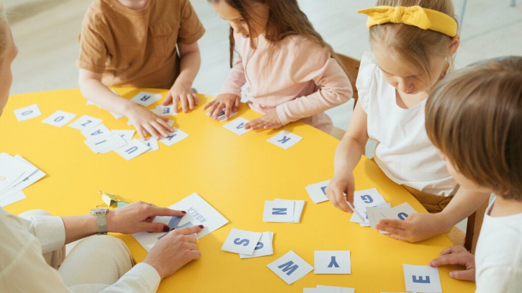 Image of a teacher working with 4 students. The students are using letters in front of them to form a word.