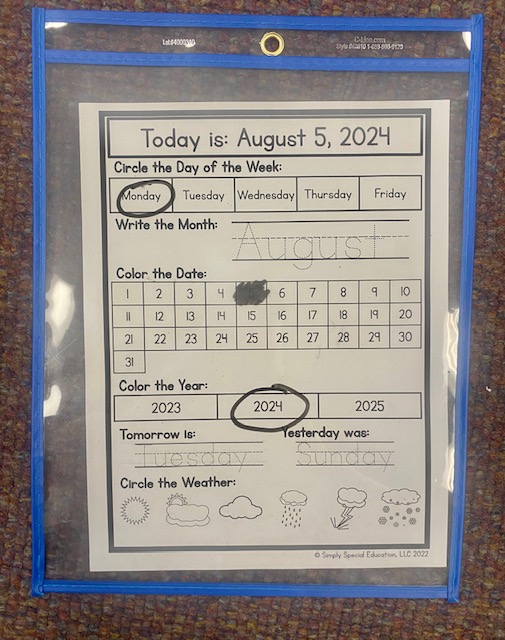 A Daily Calendar Worksheet is shown in a blue sheet protector. There are dry erase marks indicating the date.