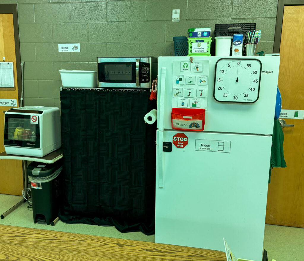 a small classroom kitchen has a refrigerator, a table top dishwasher, and a supply shelf with black curtain