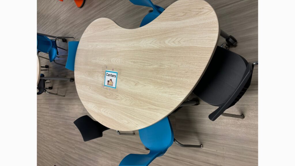 Picture of a kidney table with 5 chair around it. In the middle of the table is a blue "centers" visual.