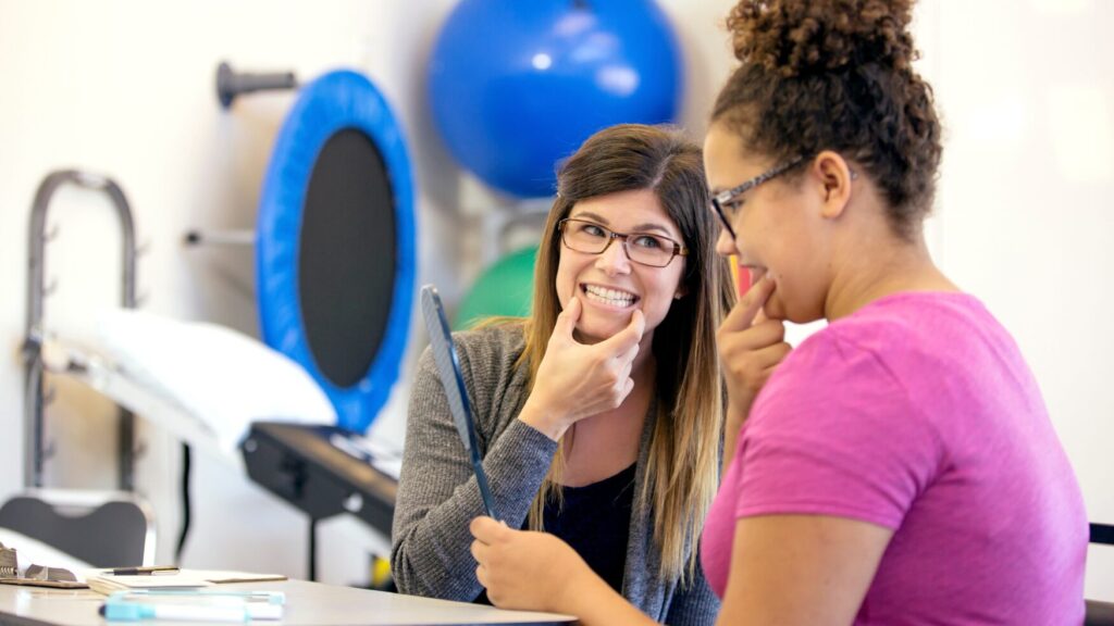a speech-language therapist assists a student by modeling a mouth shape while a student holds a mirror to view her mouth