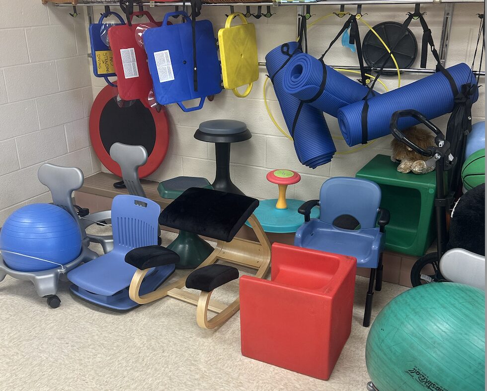seating storage in the sensory room with scooter boards and yoga mats handing on backpack hooks in the back.  cube chairs, Compass chairs, ball chairs, wobble stools, seat cushions, etc. are on the floor. 