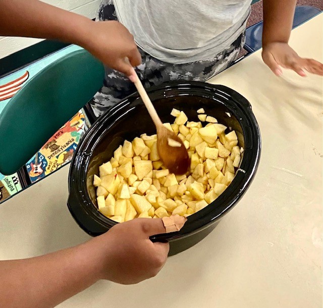 A child's arms can be seen holding a wooden spoon to stir a crockpot with chopped apples.