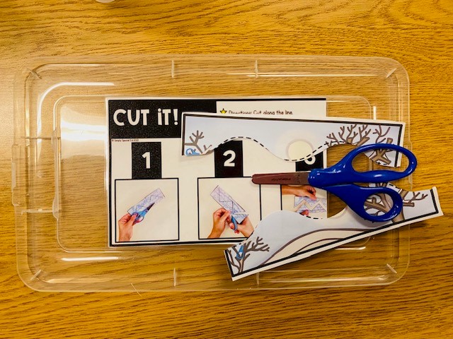 A clear plastic bin with materials for the Cut It! fine motor center, including paper pieces to be cut, a pair of scissors, and visual directions.
