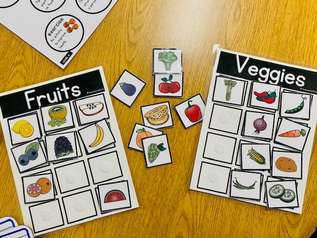 Two laminated papers labeled "fruits" and "veggies" are shown on a wooden table. There are assorted illustrated pictures of fruits and veggies sorted onto the mats.