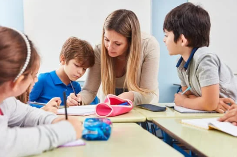 A teacher is sitting next to a student at a desk. They are writing in a notebook. Two other children are shown.