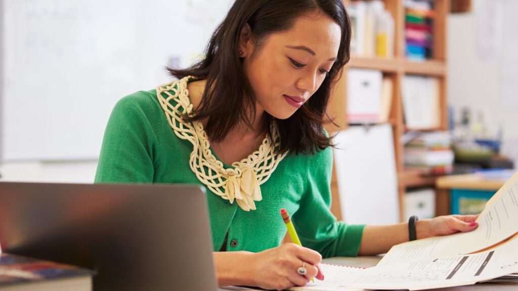 a teacher sits at her desk with a pencil in her hand writing notes in a notebook 