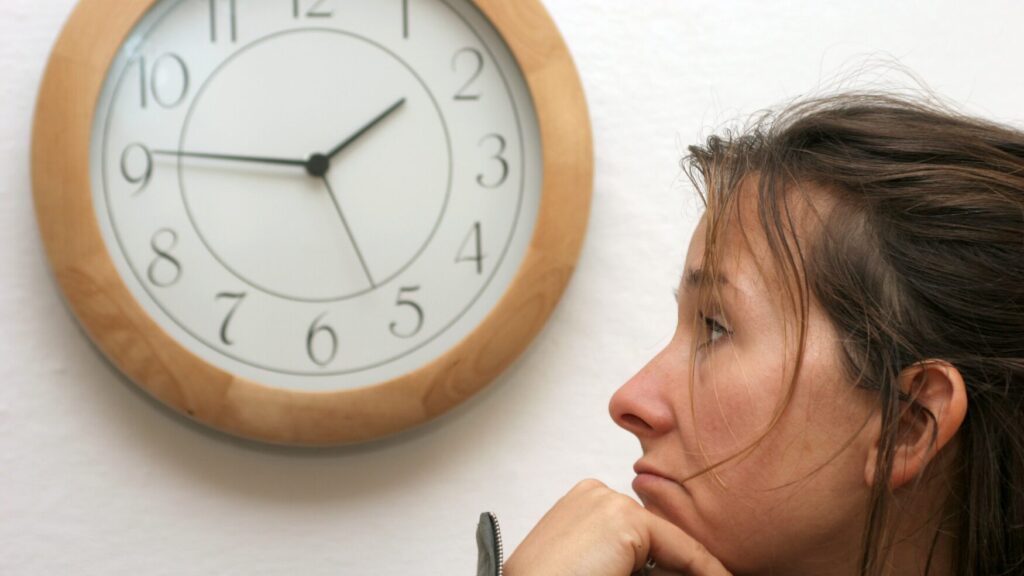 a woman waits with her hand under her chin and a clock is seen on the wall