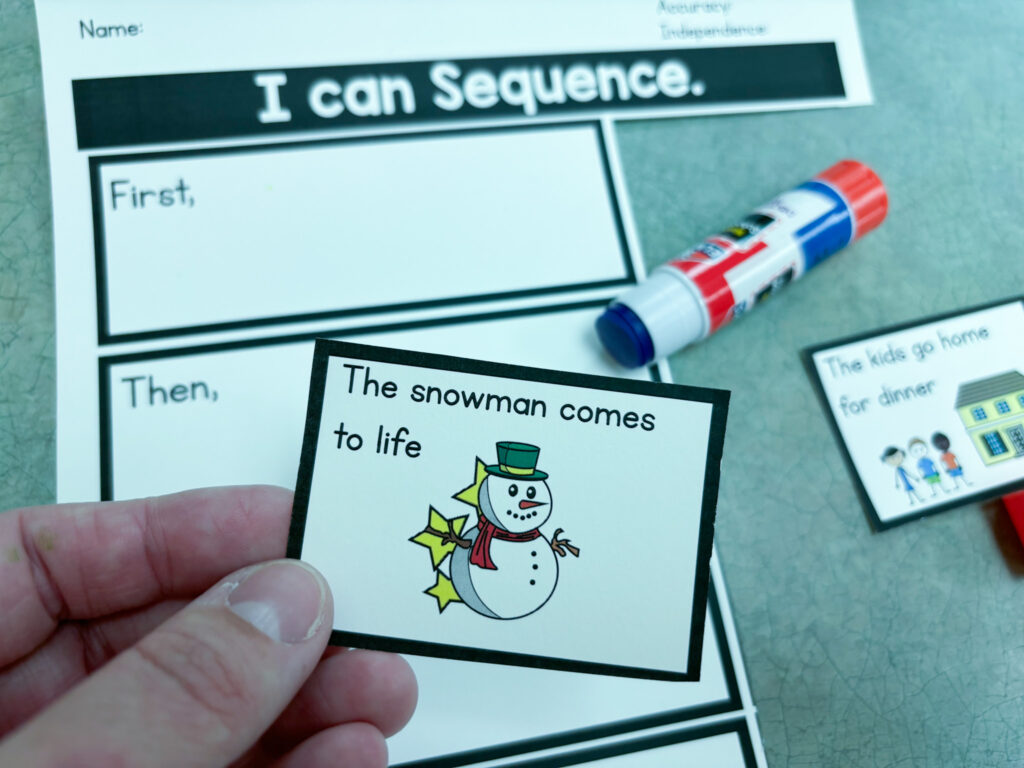 A hand holds a visual sequencing piece from a winter book companion. A paper and a glue stick sit on the table behind.