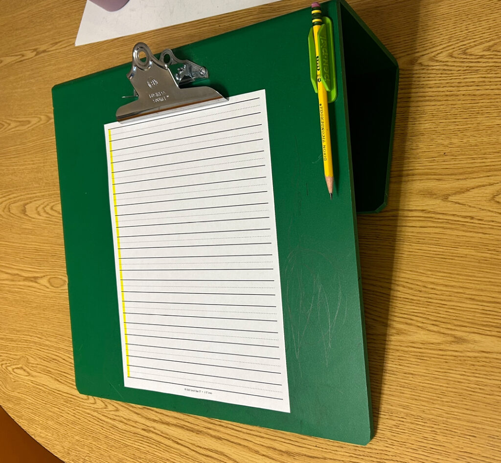 green slant board with pencil holder on the side and 3 lined paper with a left margin highlight clipped on it.