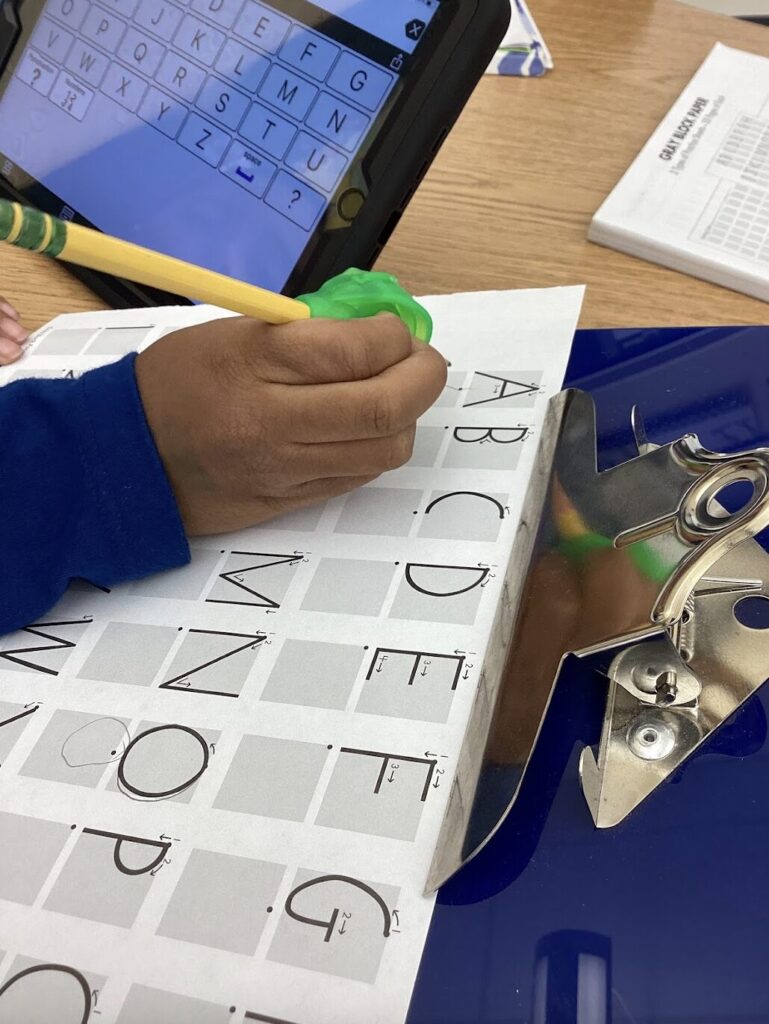 student writing on Handwriting Without Tears gray block paper with a pencil gripper on a slant board with his AAC device to say each letter