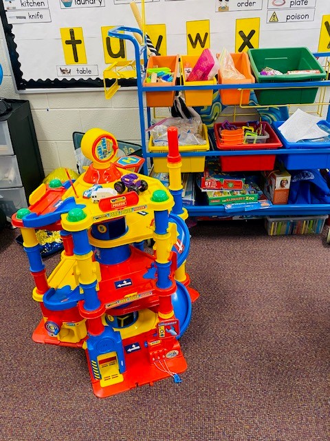 A colorful plastic toy car ramp is shown in front of toy bins in a classroom.