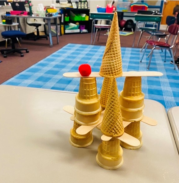 Various ice cream cones are stacked into a a pyramid, with wooden popsicle sticks in between. A red pom pom sits on top of one of the cones.