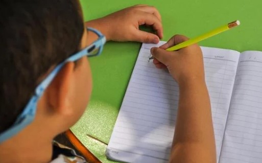A student with glasses writes in a notebook in a green table.