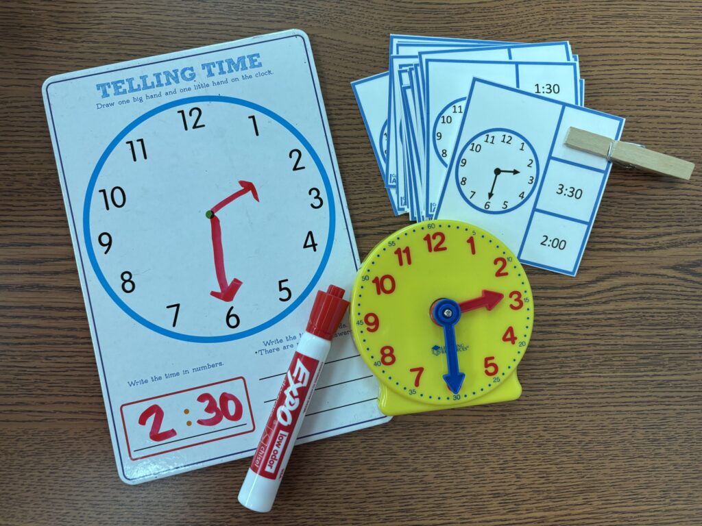 hands on tasks to teach time sit on a table including a white board and marker, clip cards, and a plastic clock with movable hands