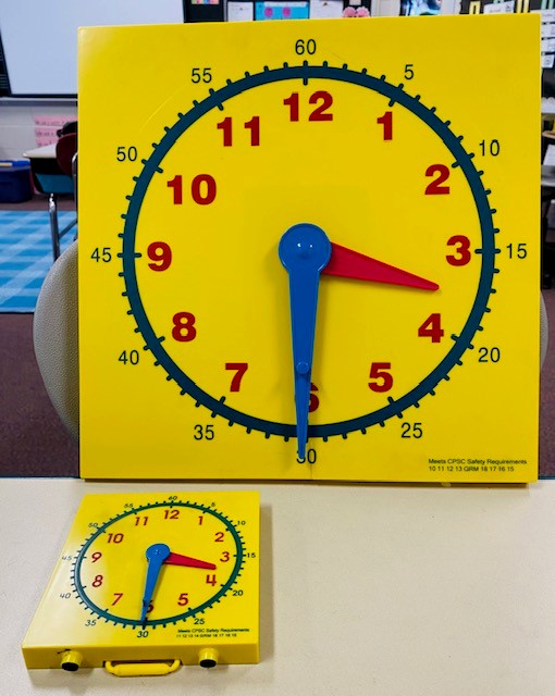 Two yellow plastic model clocks are shown. One is a large teacher-sized clock the other is a small student-sized clock. Both clocks read 3:30.