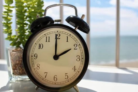 An analog clock showing 2:00 is in front of a sunny window.