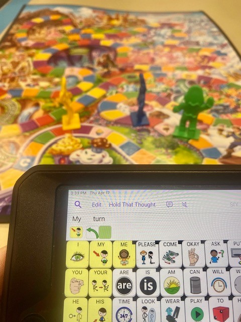 An AAC device is shown with the phrase "my turn." In the background is the board game "Candy Land"