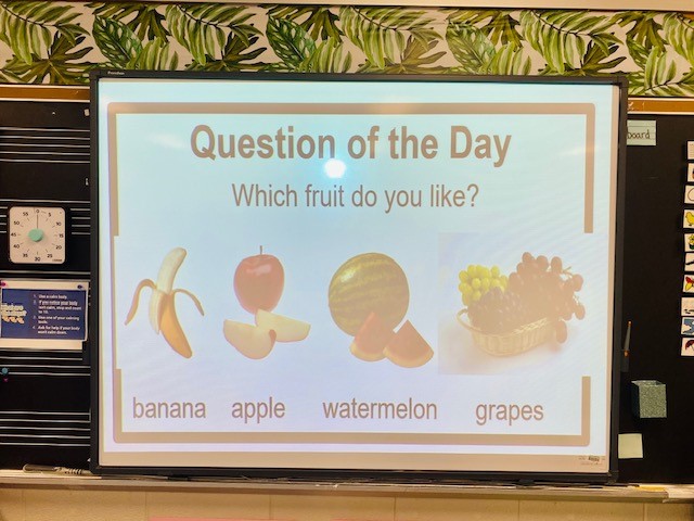A Smartboard is shown in a classroom. The slide says "Question of the Day" and asks "Which fruit do you like?" with choices for banana, apple, watermelon, and grapes.