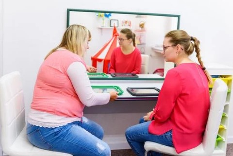 Two women sit at a table facing each other. They each have a tablet in front of themselves.