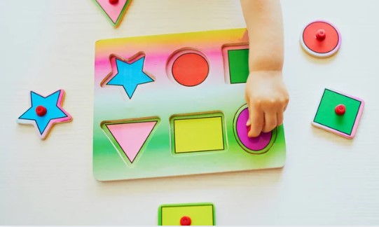 A green wooden shape puzzle is shown on a table. A child's hand is reaching for one for the puzzle pieces.