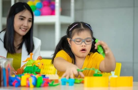 A young girl in a yellow shirt plays with toys at a table, while a teacher sits close behind her watching.