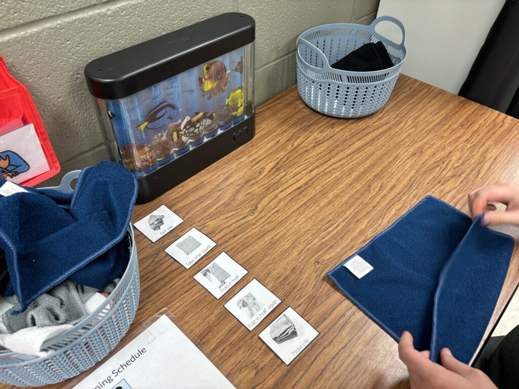a functional life skills task box is shown that requires a student to fold hand towels. visual task analysis is placed on the desk to show student step by step how to complete the task.