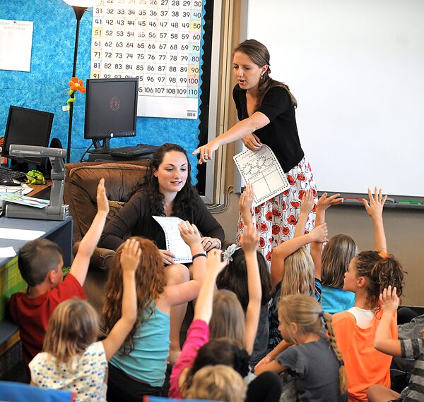 Two teachers teaching at the same time, one calls on a student with their hand up while the other one watches along 