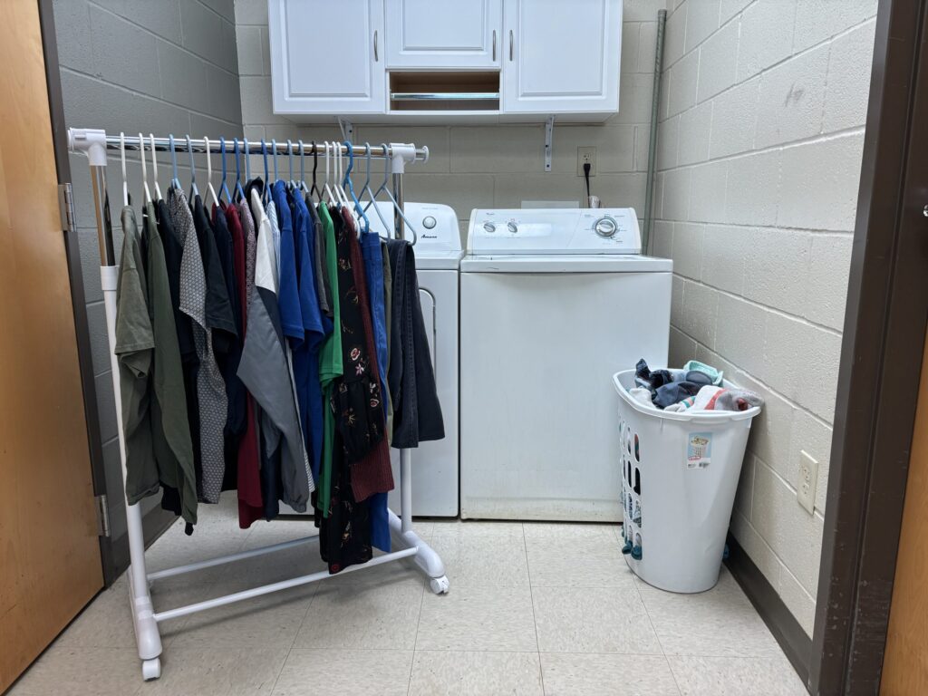 clothing is hung on a rolling rack near a washer and dryer and a basket of dirty clothes placed for students to work on life skills