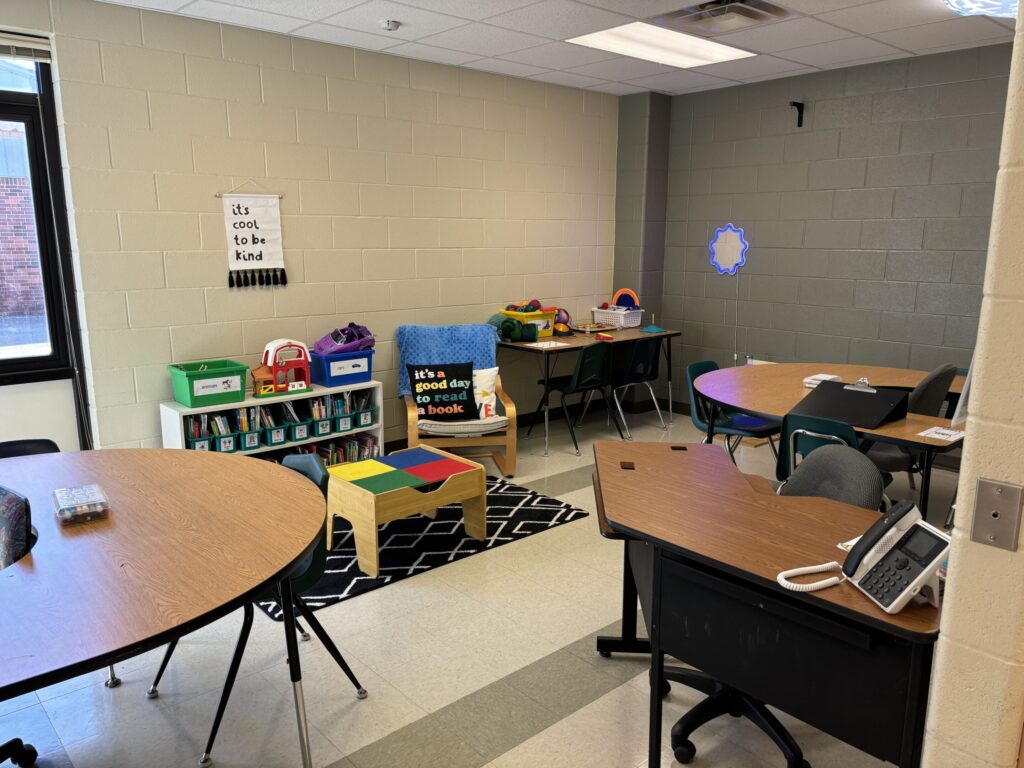 a variety of tables in shown in a classroom, including an adult desk, small group tables, a sensory table, and a bookshelf of student free choices