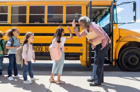 A line of students waits to board a yellow school bus. A teacher s giving a high-five to the first girl in the line.