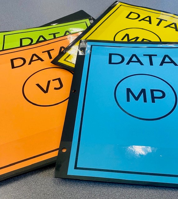 Four folders are on a table. The folders have colorful labels in green, blue, orange, and yellow. Each folder is titled "DATA" and has student initials.