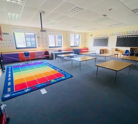 An elementary school classroom is shown. there is a colorful area rug in the front of the room, six wooden tables, and two black bulletin boards in the background.