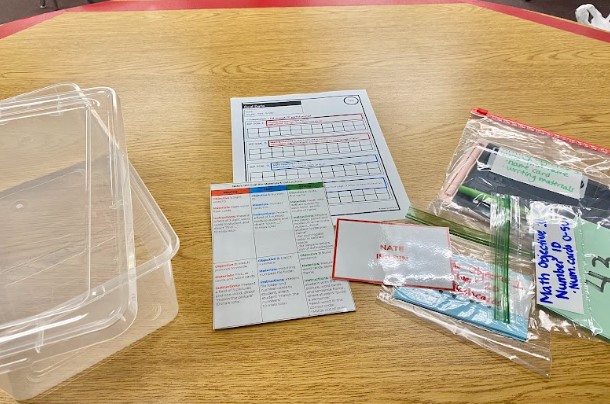 An IEP Goal bin is spread out on a table. THe clear pastic bin is shown with data sheets and bags of materials.