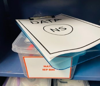 An IEP Goal Bin is shown on a shelf, with a data clipboard on top.