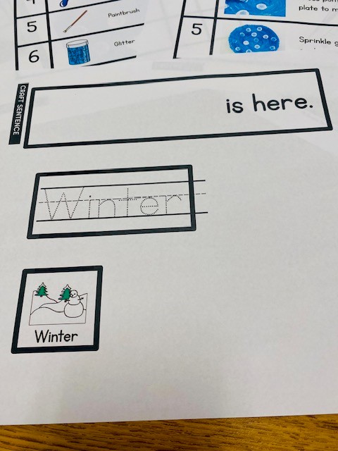A printed paper with a sentence reading "__________ is here." Under the sentence are response choices including a traceable version of the word "winter," and a "winter" PEC image.