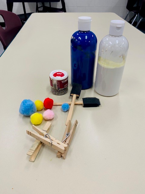 Art supplies including apint, paint brushes, glitter, pom poms, and clothespins are shown on a table in a classroom.