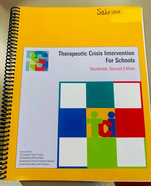 The Therapeutic Crisis Intervention for Schools workbook is shown It is a yellow spiral-bound book with a colorful "tci" logo.