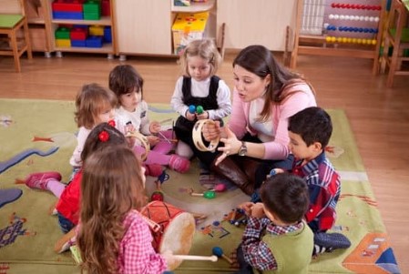 In a classroom, a teacher sits with students in a circle on the floor. They are holding toy instruments.