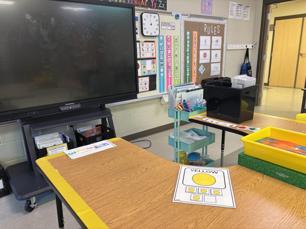 a classroom group table is edged in yellow ductape to signify the yellow station. Yellow visual icons are placed on the table for student check in.