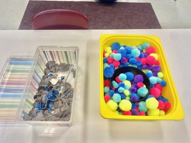 Two sensory bins sit on a table in a classroom. One contains kinetic sand and one contains colorful pom poms.
