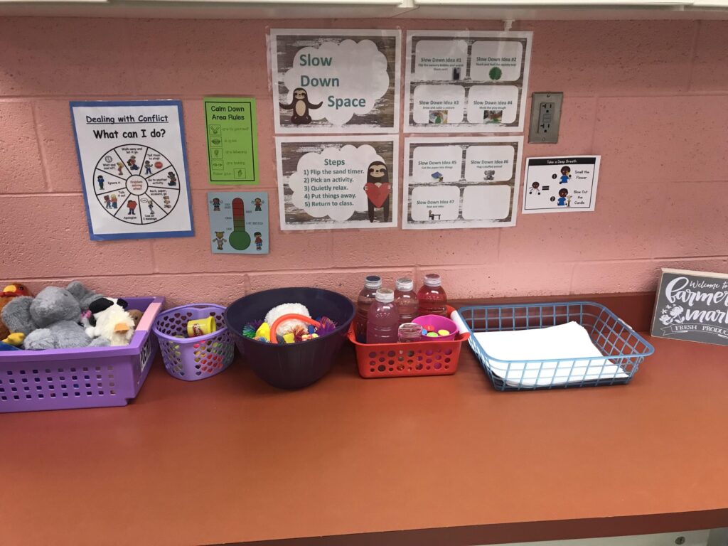 A table in a classroom with posters labeled "slow down space" with a sloth and other posters that describe how to deal with conflict, along with baskets of sensory toys and stuffed animals