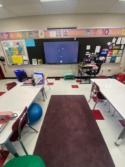 An image shows part of a classroom with two white tables facing a smart board