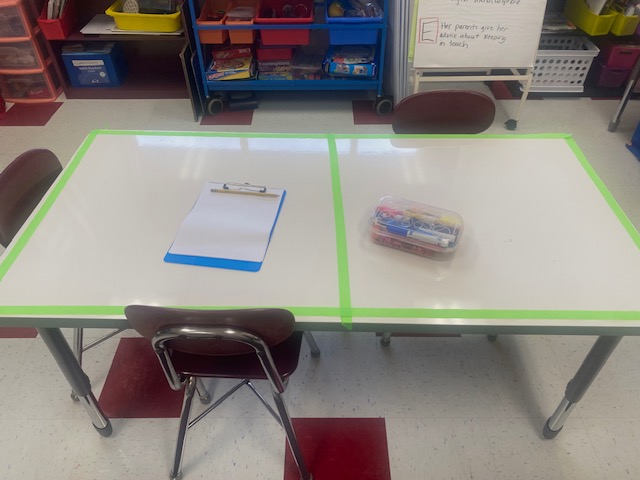 A white table in a classroom has green masking tape around its edges and down the middle, dividing it into two parts.