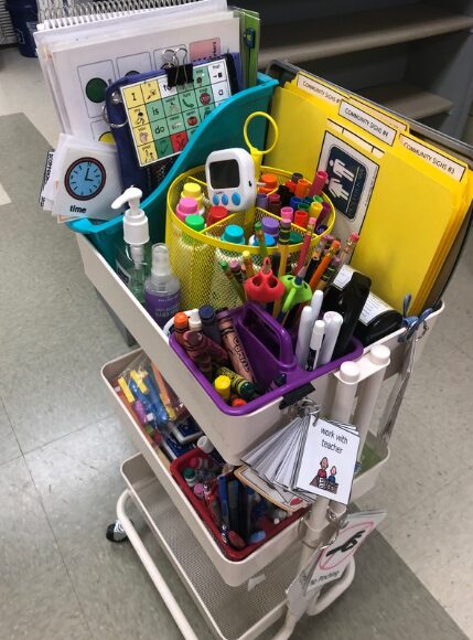 A rolling cart is stocked full of supplies including visuals, timers, various writing utensils, math tools, and hand sanitizer.