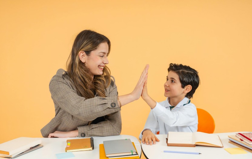 A teacher and student high-five at a table. There are notebooks and pens on the table.