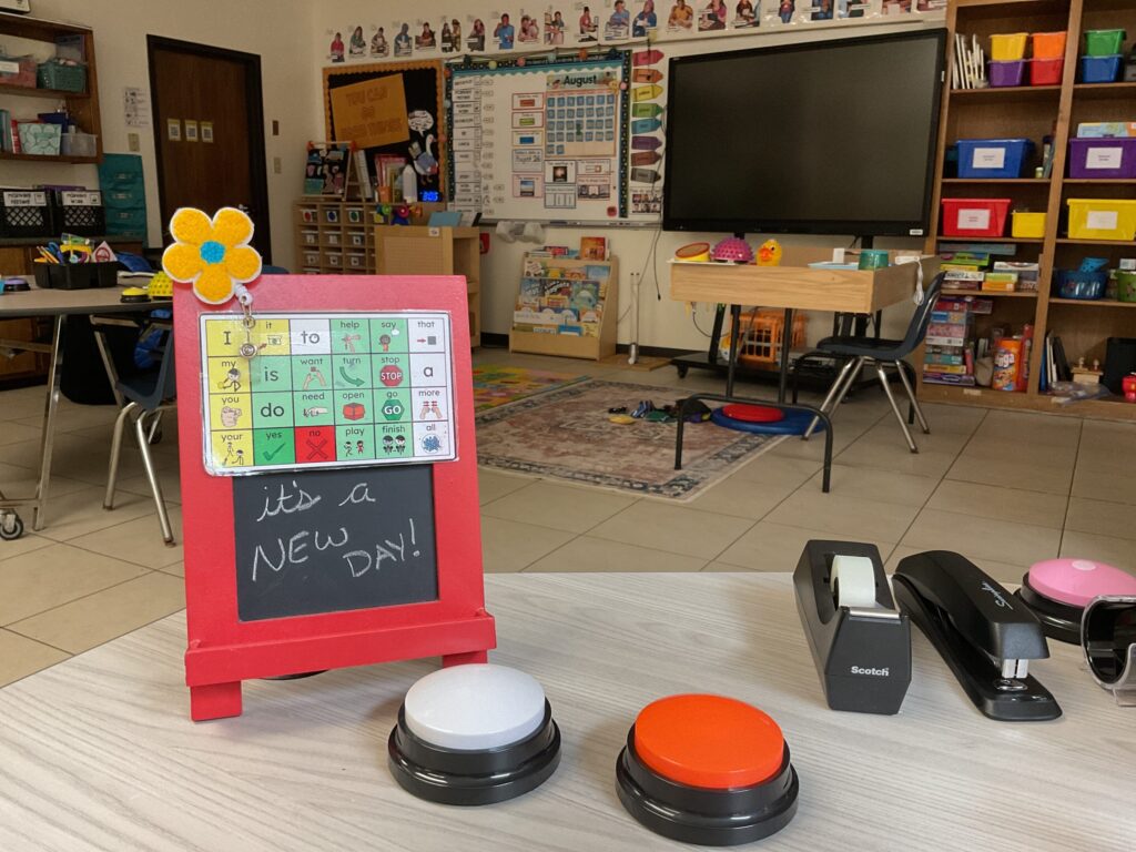A chalkboard atop a teacher’s desk reads, “it’s a new day!” . The photo overlooks the rest of the classroom, shelves, and board.