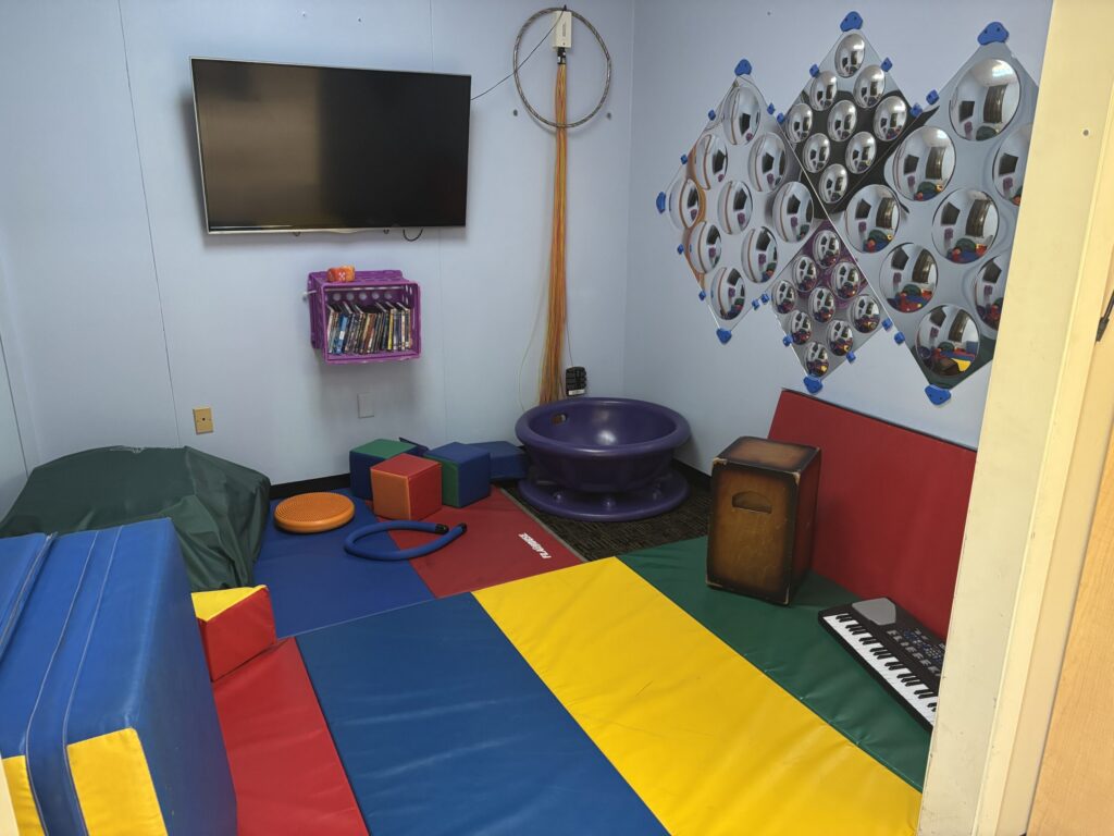 A sensory room is covered with colorful mats and contains musical instruments, a crash pad, and mirrors on the wall.